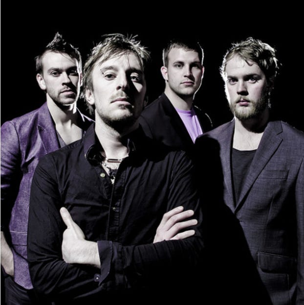 A very slick 00s looking band of four white dudes in smart clothes. they look like they wish they were MUSE