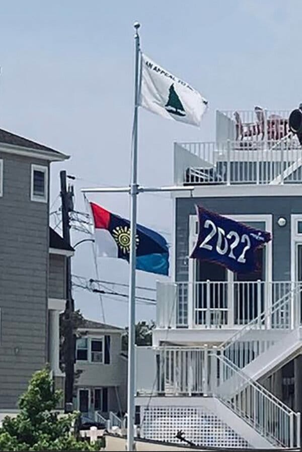 A flagpole displaying a white flag with a pine tree and the phrase “An Appeal to Heaven,” a blue “2022” flag and a multicolored flag with a yellow sun in the center.