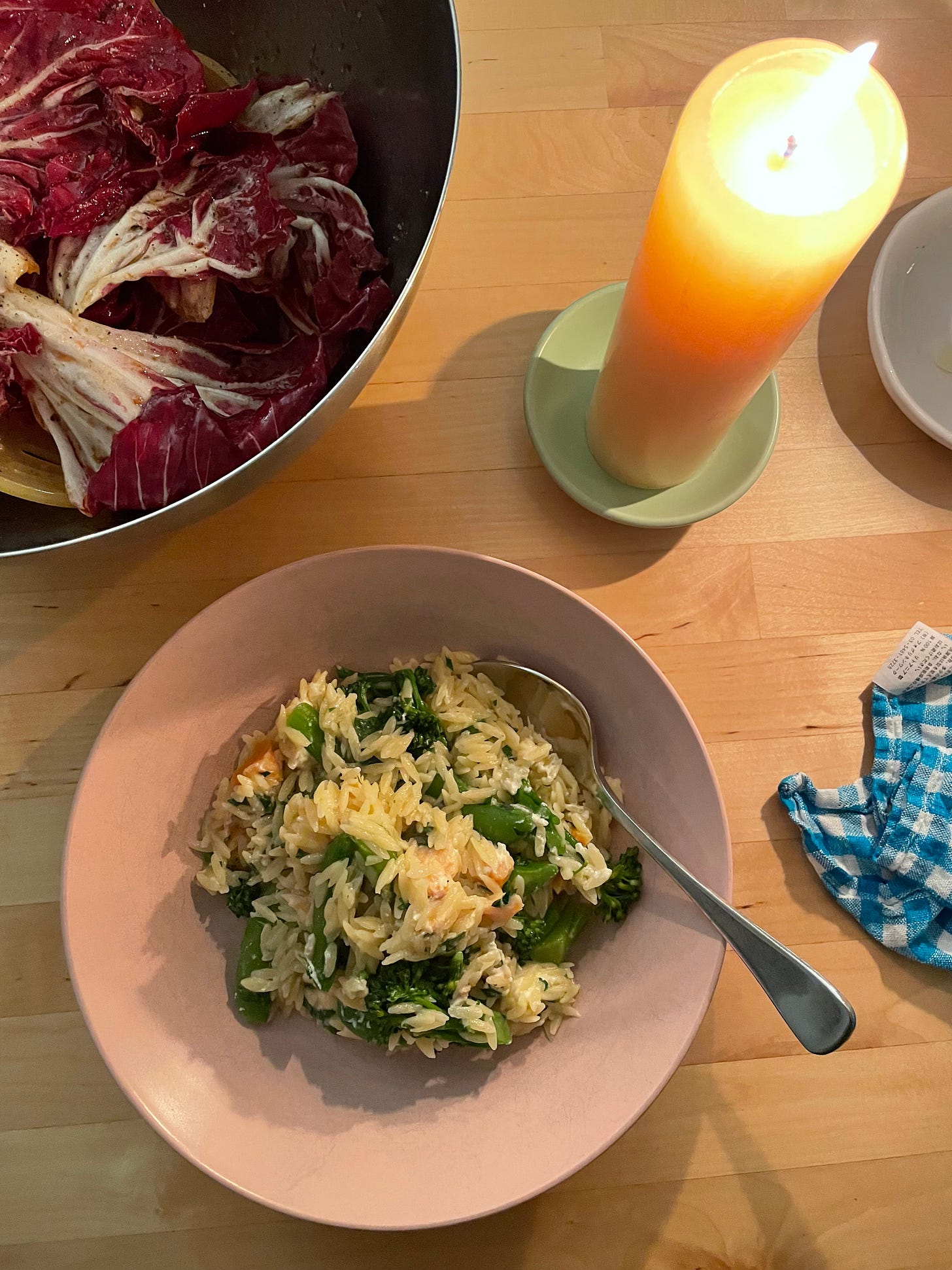 A bowl of cold-smoked salmon risoni with baby spinach. A column candle lights the dinner scene. A blue and white chequered napkin nearby. The meal is accompanied by a big bowl of raddichio salad topped with lots of pepper, olive oil and balsamic vinegar.