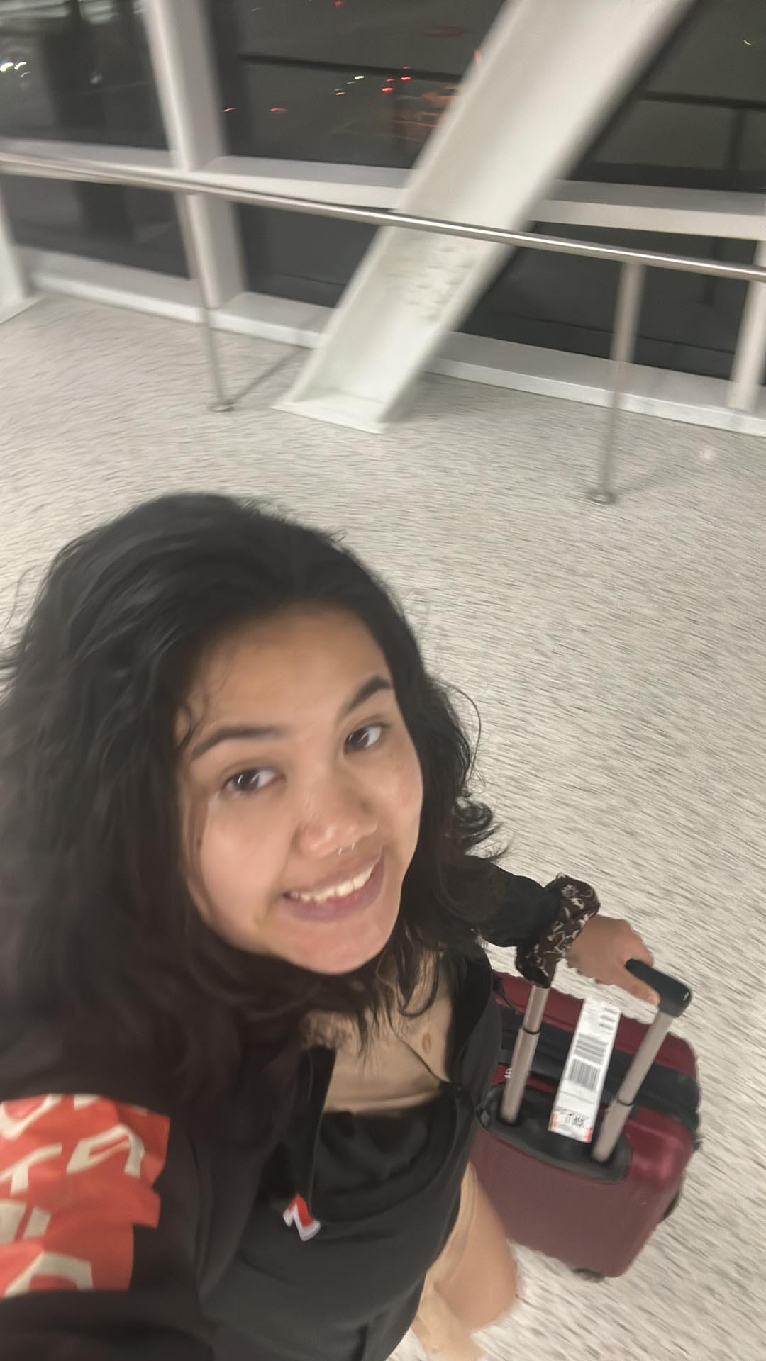 cami (young 21 year old woman with messy brown hair) grinning anxiously at the camera with her luggage