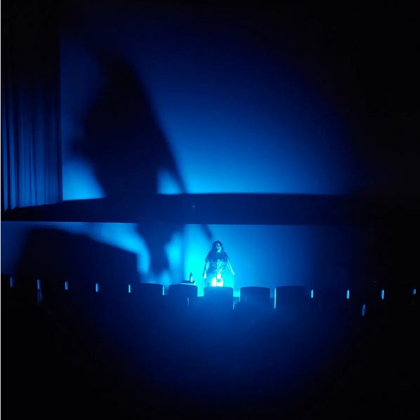 Rebekah Del Rio in near-total darkness, only illuminated by a blue light in front of her and casting a large shadow on the screen behind her, swaying and singing into a lone microphone with no accompanying band or musicians