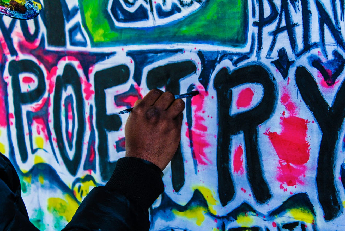 A hand reaches out to a wall brightly graffitied with the word Poetry.