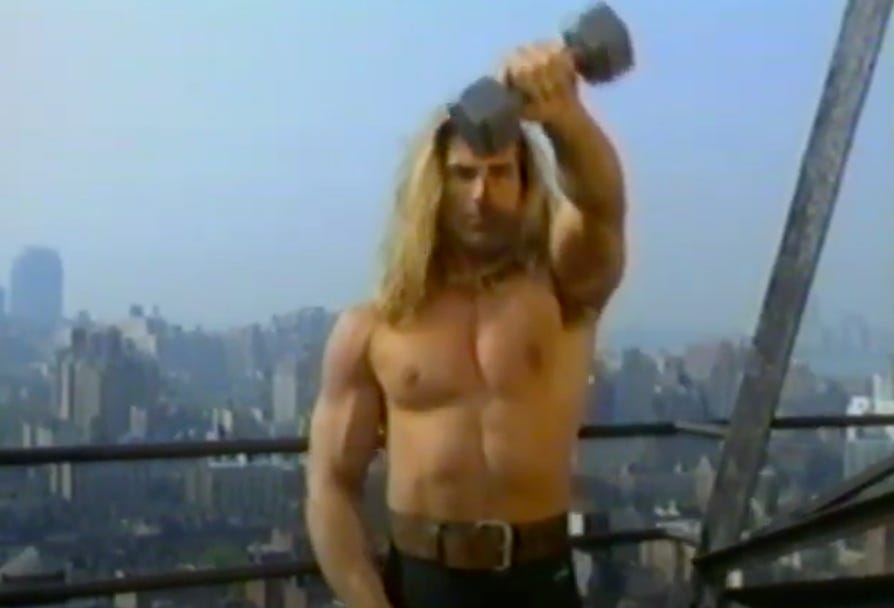 This is a screenshot from Fabio Fitness: Fabio is working out alone on top of a New York City skyscraper. He is lifting a weight over his head and looking at the camera. He is wearing a large belt but no shirt. 