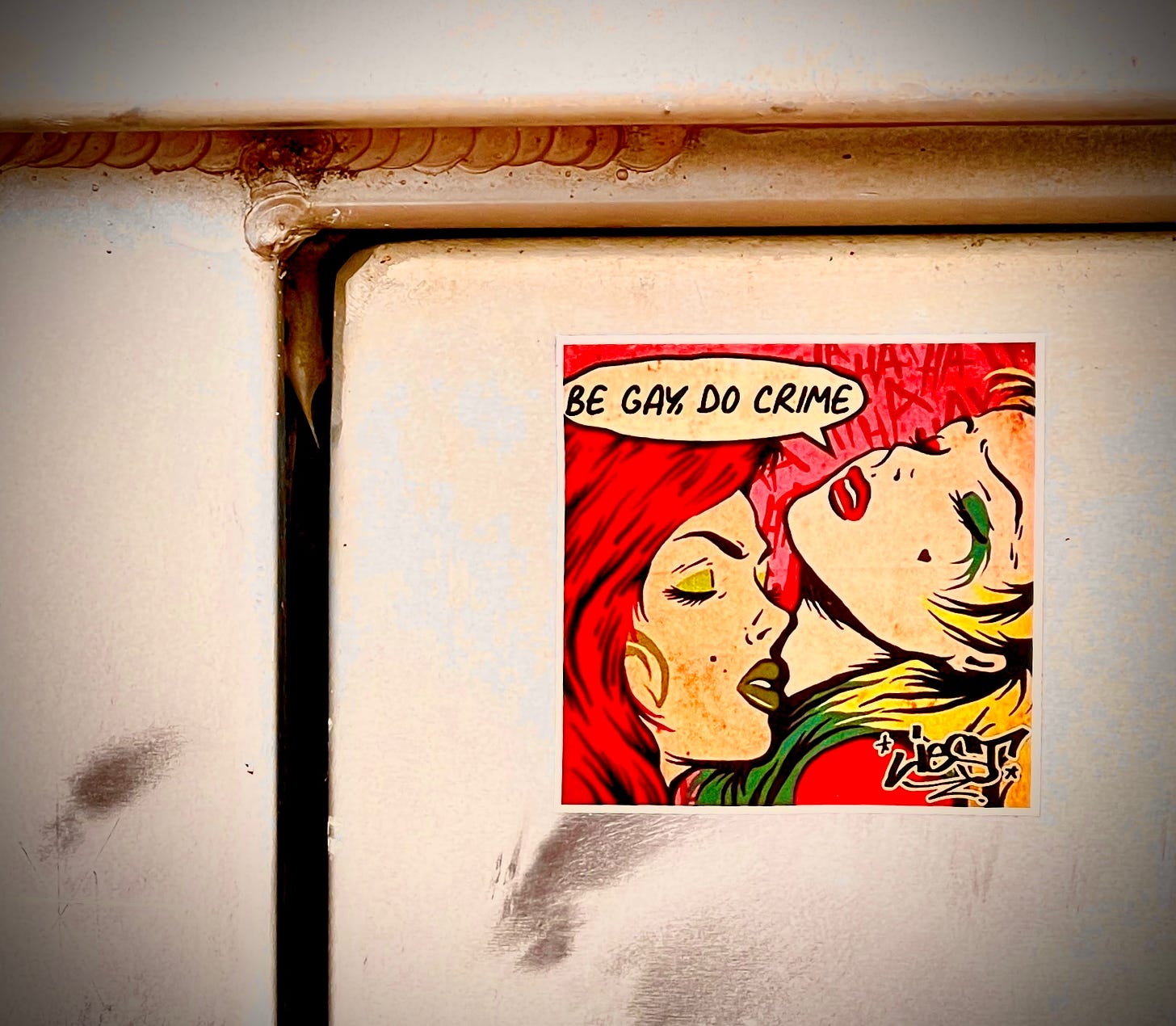 Colorful sticker on an indistinct gray metal background showing two passionate woman with bold lipstick and eye shadow, and dyed hair, with their eyes closed looking and a text bubble showing one of them “Be gay, do crime”.