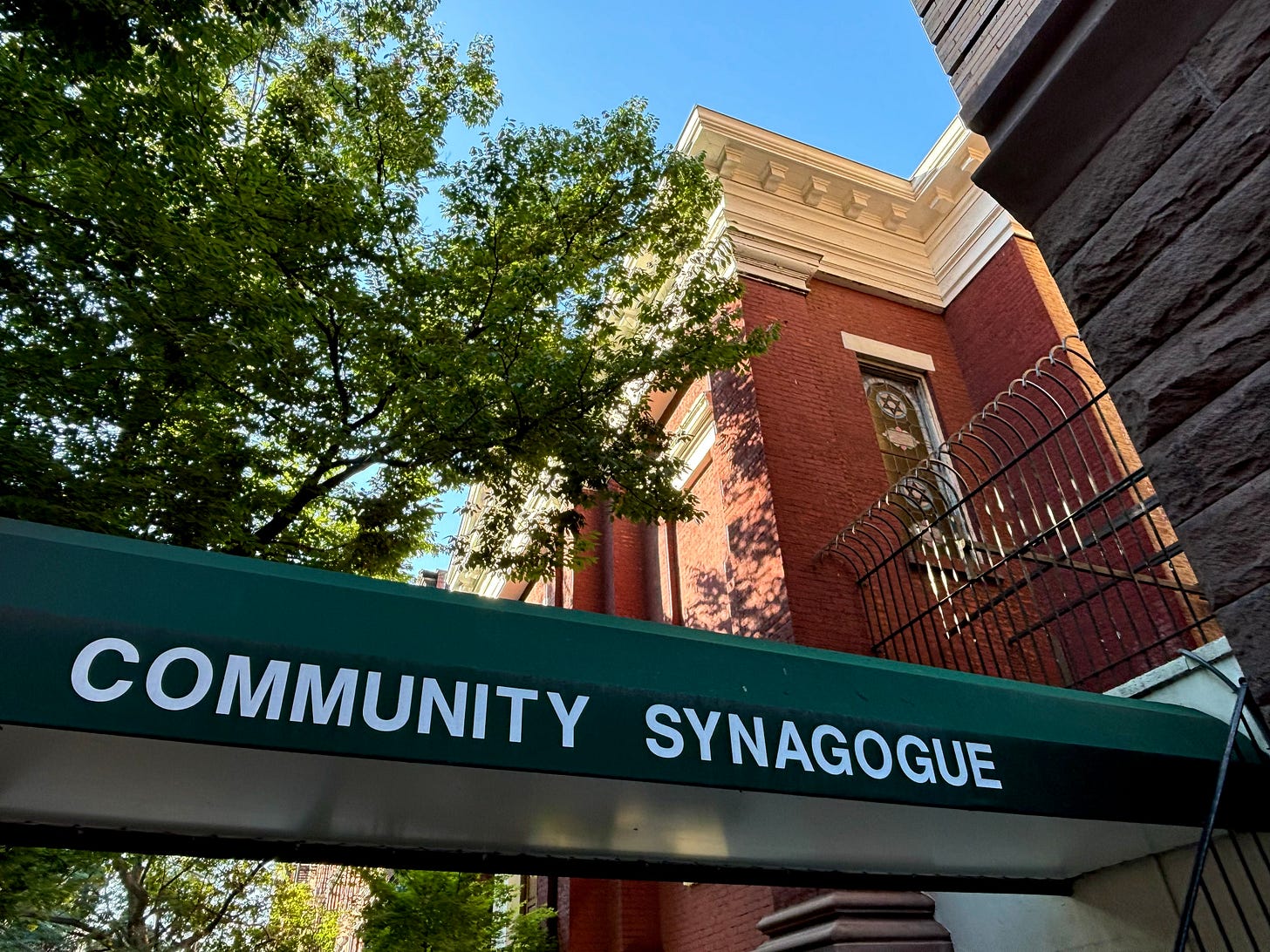 The green awning of the synagogue on 6th Street. It says COMMUNITY SYNAGOGUE in white letters. In the wall behind is a stained glass window in the shape of a Star of David. 