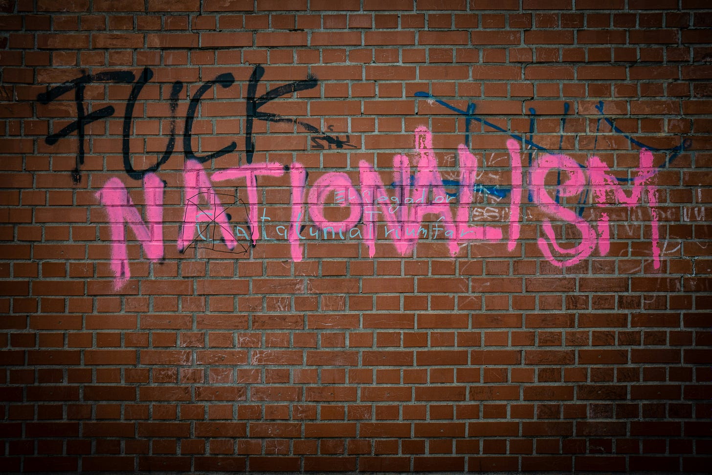 The phrase "fuck nationalism" - "FUCK" is painted in black and "NATIONALISM" in pink. Painted on a red brick wall, with some other smaller graffiti showing as well.