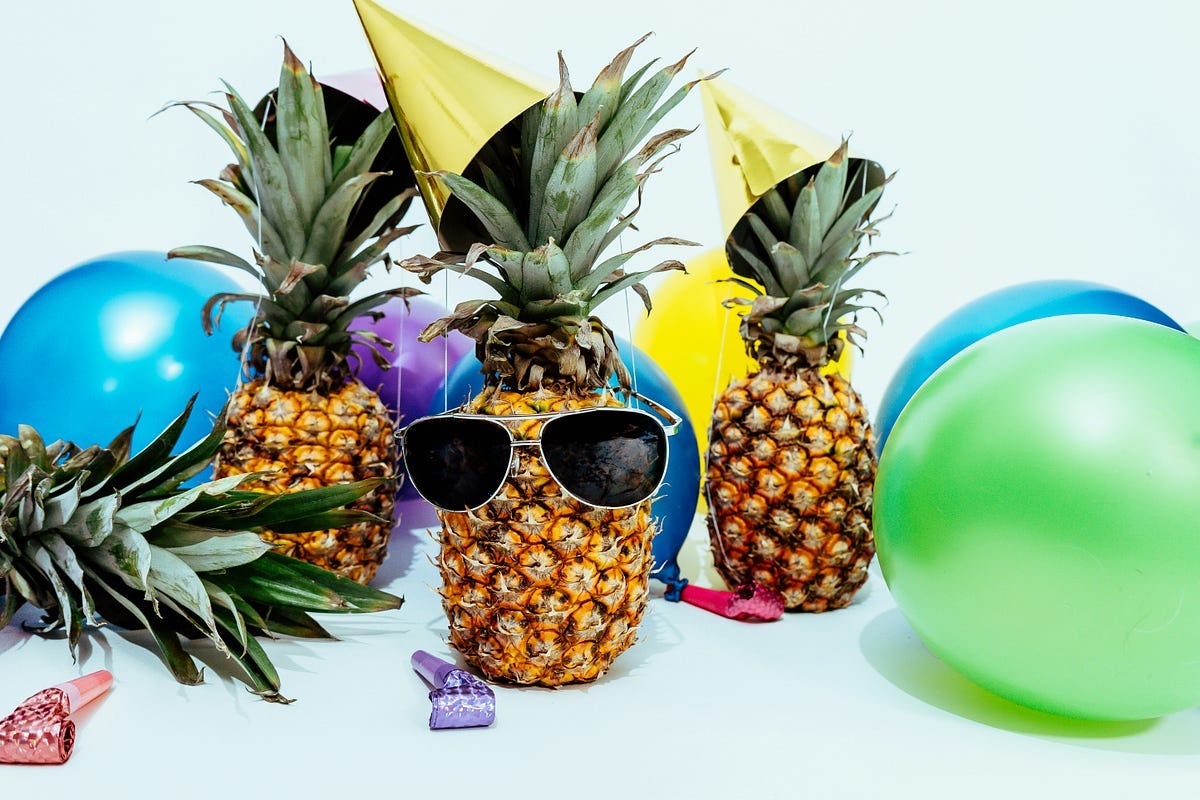 a pineapple with sunglasses and party hat. Surrounded by baloons, in celebration