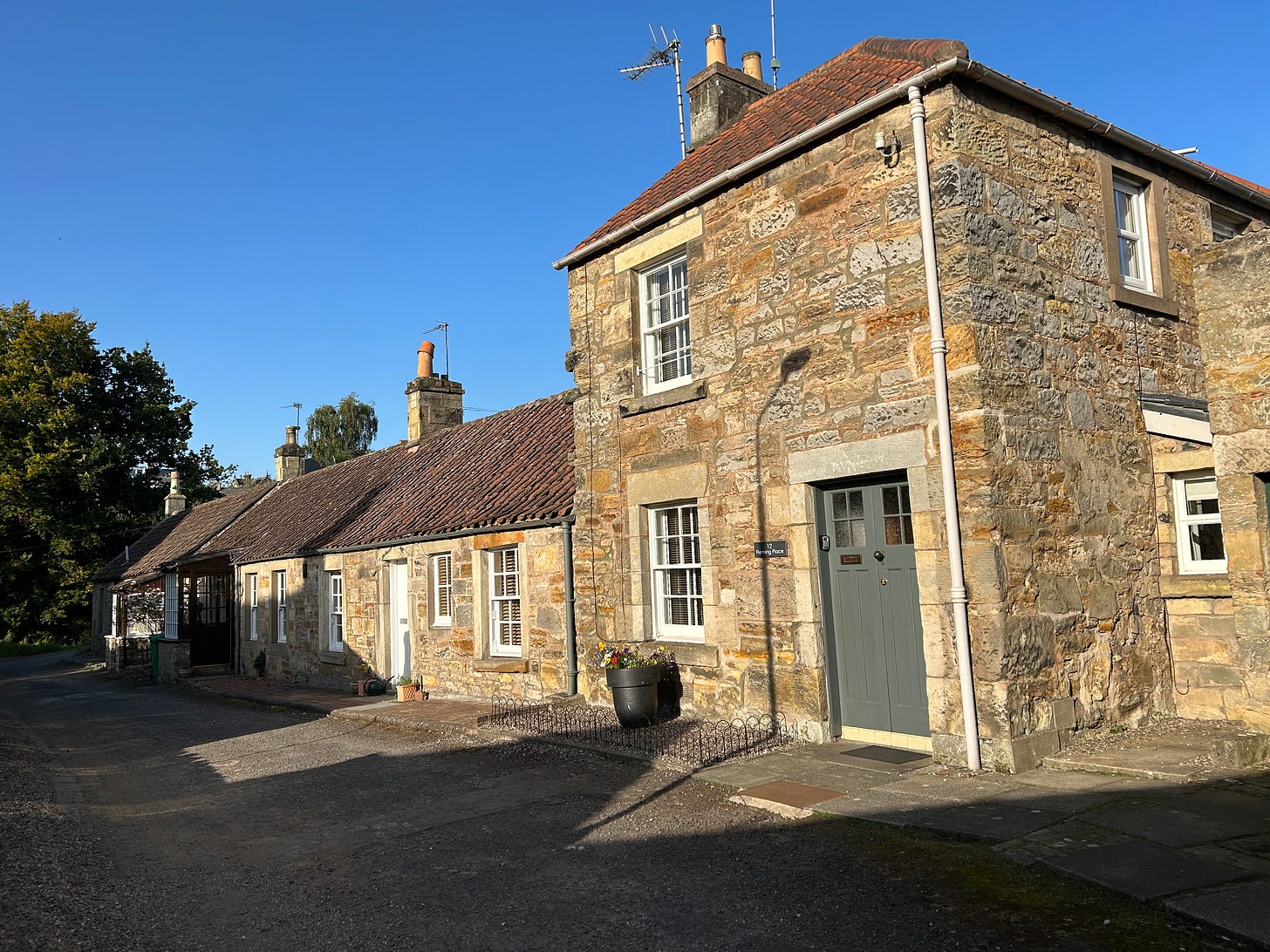 A two-level stone cottage, built with yellow and light-brown stones, sits at the end of a stretch of one-level cottages. The structures have red-brown roofs and cheerful white-paned windows. 