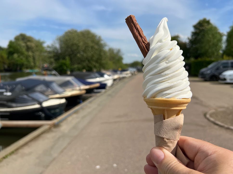 A hand holds a vanilla soft-serve ice cream cone with a chocolate flake inserted in it, known as a “99,” in front of a riverbank lined with moored boats. The background is slightly out of focus, showing a tree-lined waterside path under a blue sky with scattered clouds.