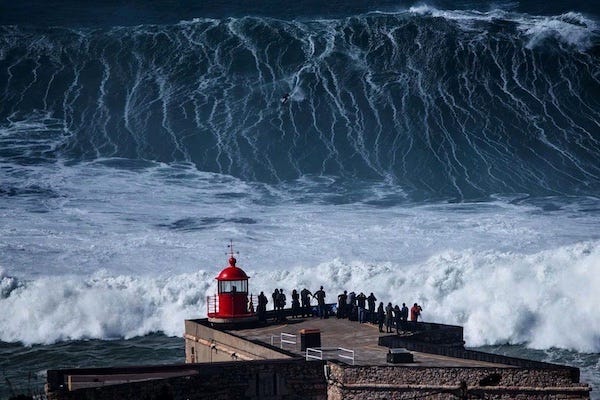 Photograph (Credit: Red Bull) of a terrifyingly large wave -- no, bigger than the one you're imagining. About ten times bigger, as seen from a vantage above a building on the shore. There are some spectators standing on the roof of the building looking at the insanely powerful wave and the teeny tiny person on a surfboard riding down its face.