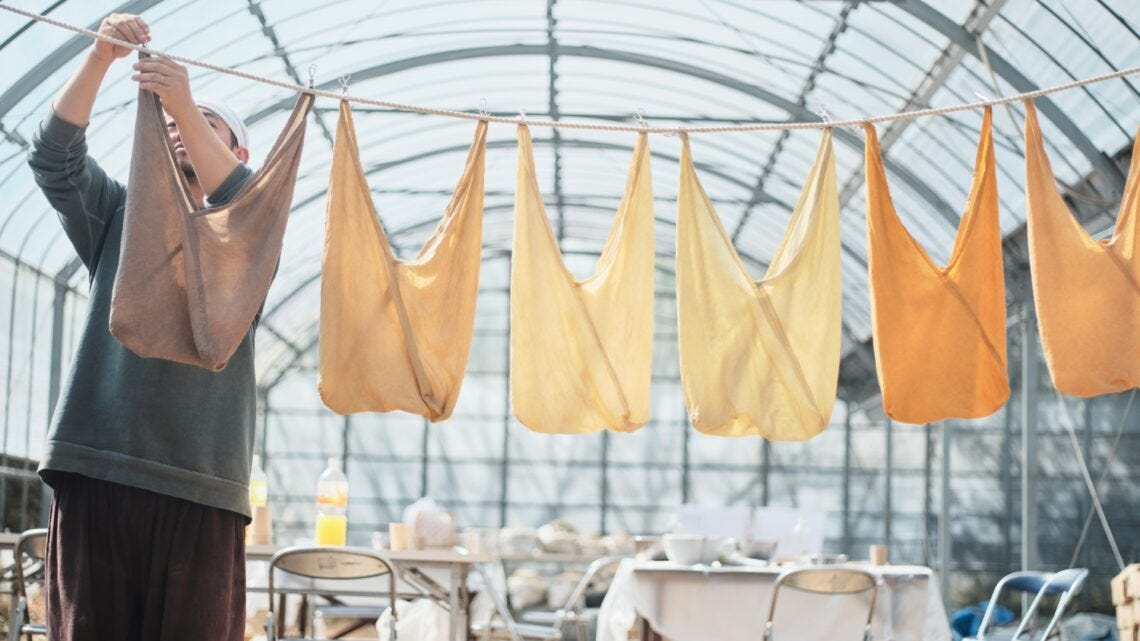 An artist hangs cloth bags of various light earthy tones on a rope line in a large greenhouse.