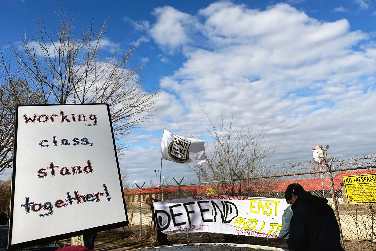 on the left, a white poster with black writing and red and blue outlines reads "working class, stand together!" in the background people hold up a white banner with red and black and yellow words reading "DEFEND EAST PHILLIPS" "no demolition" a flag flies in the background reading 'Climate justice committee" and the Roof Depot building and water tower in the background behind a tall barbed fence with a yellow no trespassing sign