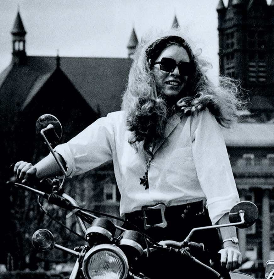 A woman with sunglasses and long, wild blonde hair sits smiling on a motorcycle.