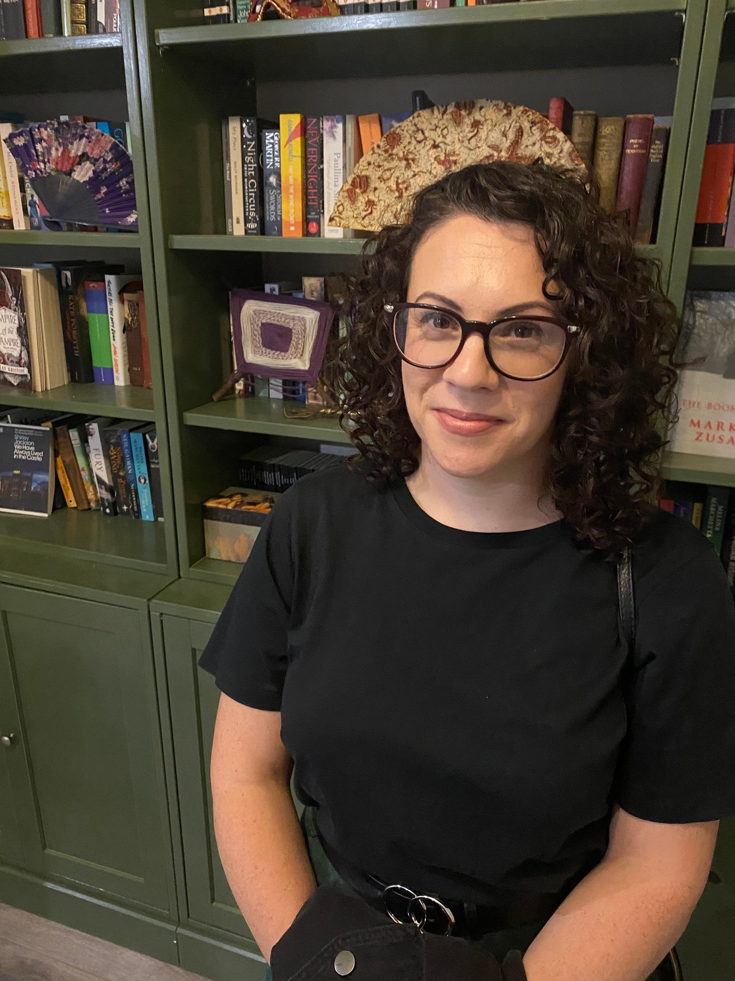 A picture of the author standing in front of a bookcase messily arranged with books and trinkets. The author has glasses on, curly hair, an awkward smile, and a black t-shirt.