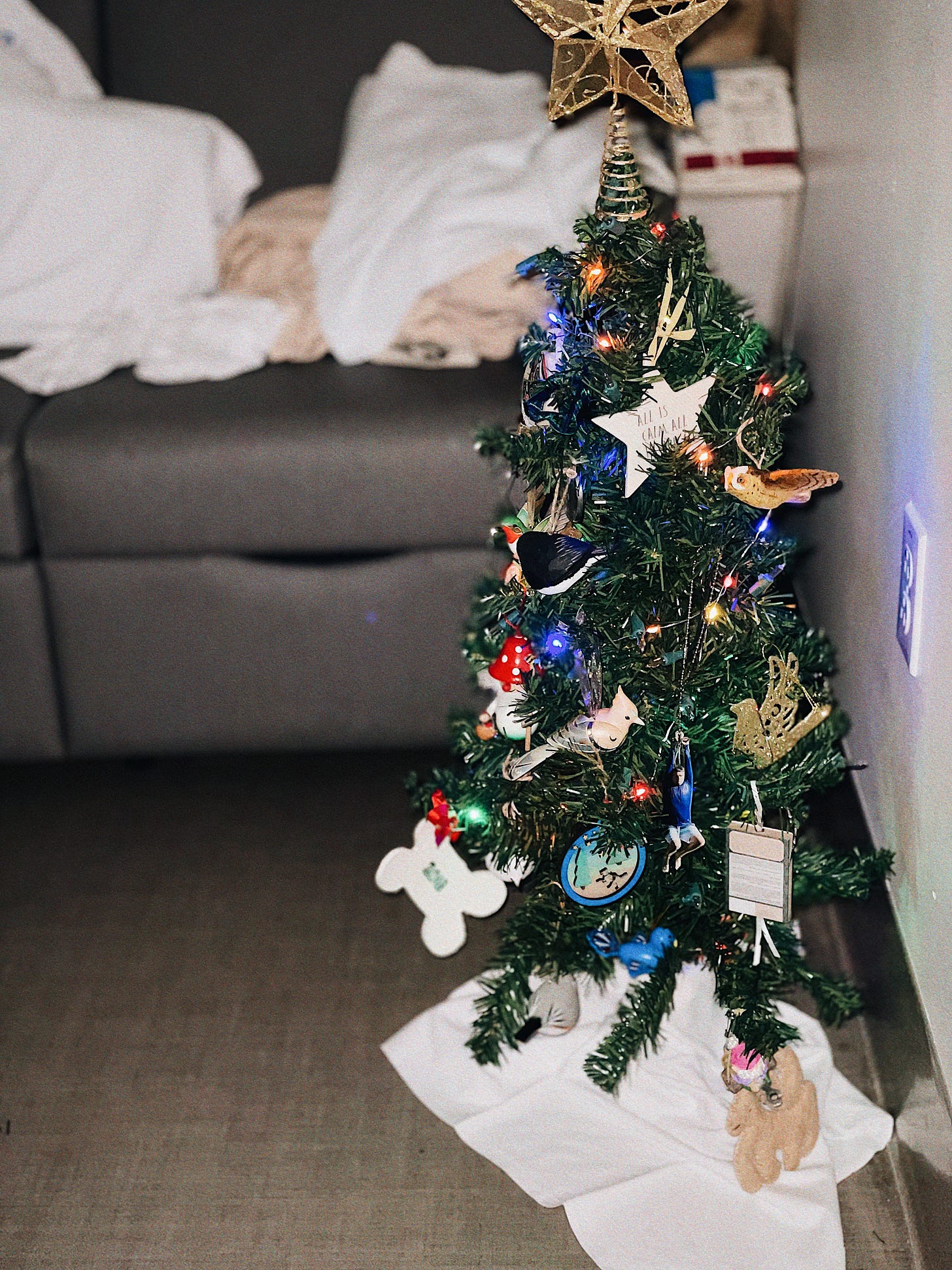 Alt-text: Small decorated Christmas tree with colorful lights, ornaments, and a gold star topper, set on a white tree skirt near a gray couch.