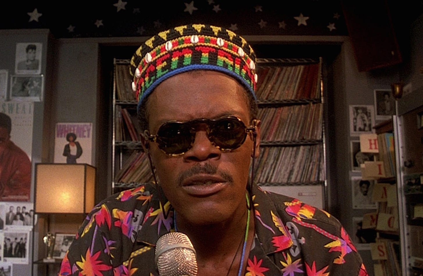 This image shows Mister Señor Love Daddy (played by Samuel L. Jackson, credited as Sam Jackson), disc jockey for Do the Right Thing's fictional radio station, WE LOVE 108FM, staring directly into the camera as he rebukes five of the film's characters immediately after they engage in what the movie's screenplay calls the "Racial Slur montage." Love Daddy wears sunglasses and a colorful kaffiyeh hat while sitting in front of bookcases full of LP albums.