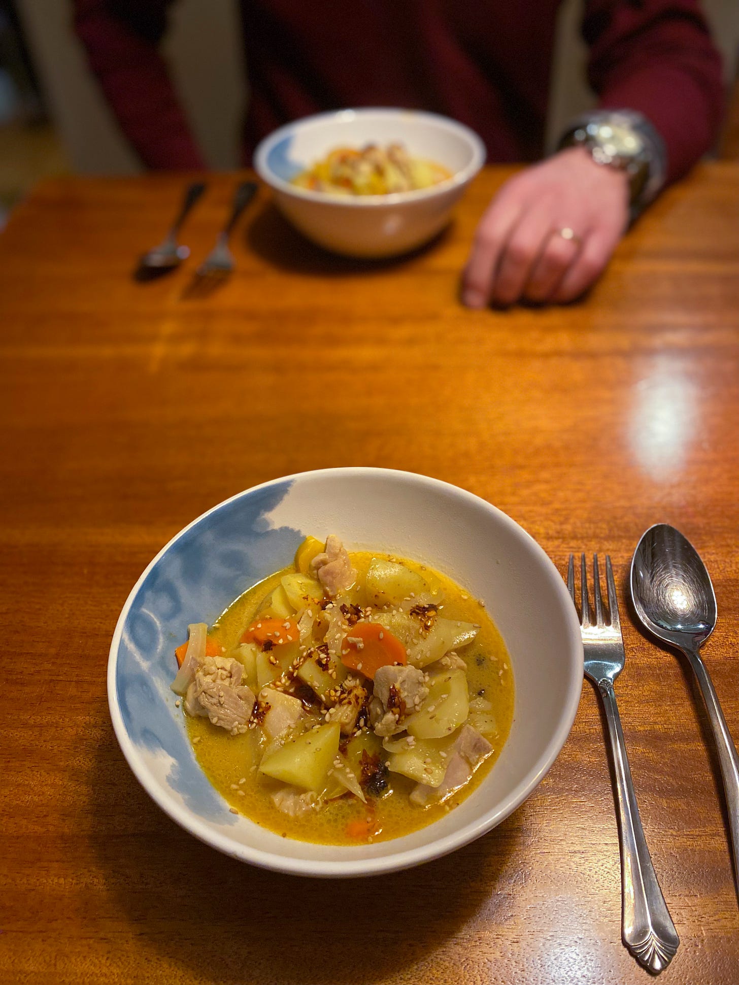 Two white and blue bowls of yellow curry with chicken, potatoes, carrots, & peppers across from each other on the table. The curry is topped with sesame seeds and chili oil, and next to each bowl are a fork and spoon. Jeff's hand rests next to his bowl across the table.