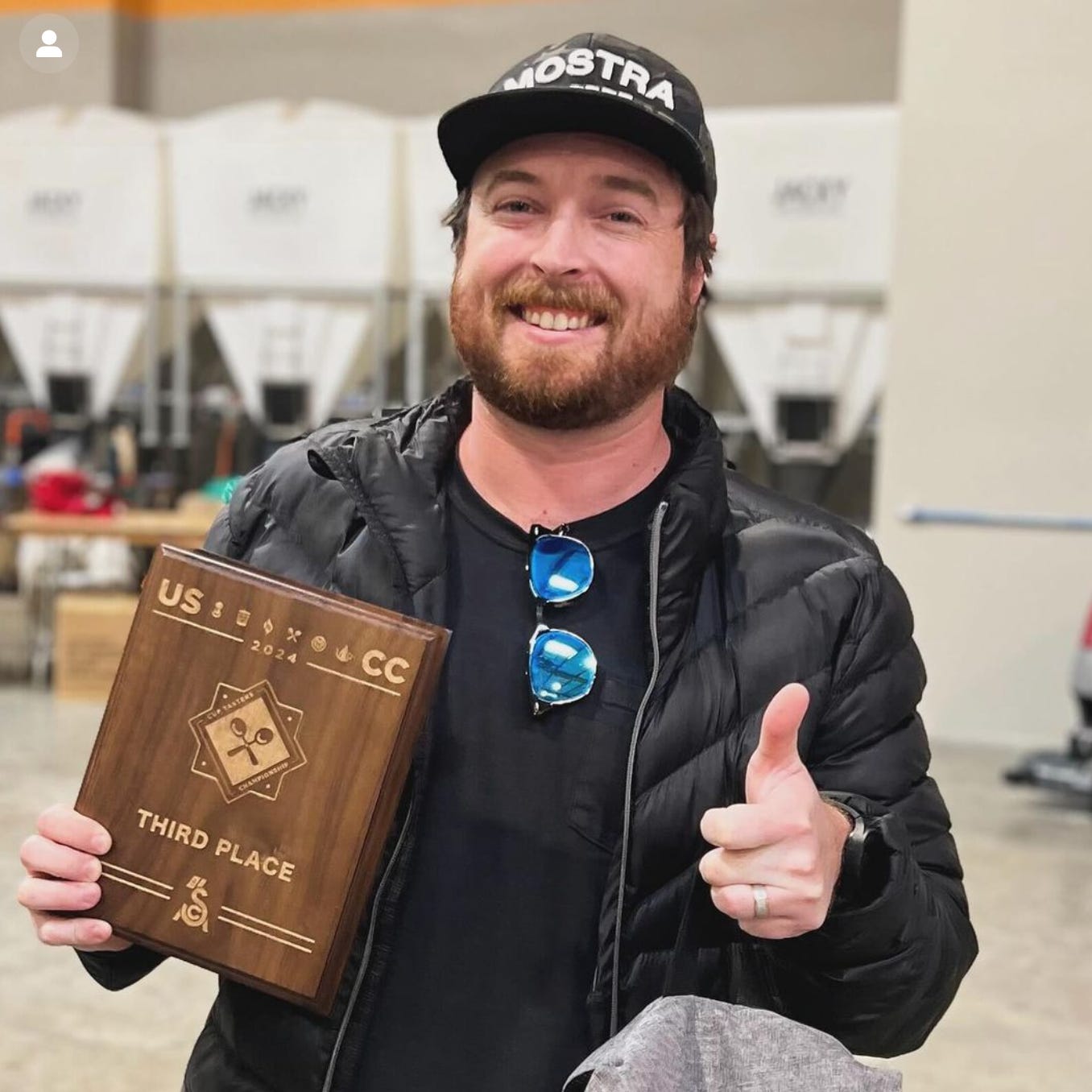 A bearded white man holds a third place plaque and flashes a smile and a thumbs up at the camera. He's wearing a black tee under a black puffy coat, sunglasses tucked into the collar of his shirt, and a camo Mostra Coffee cap. Coffee equipment is blurred in the background.