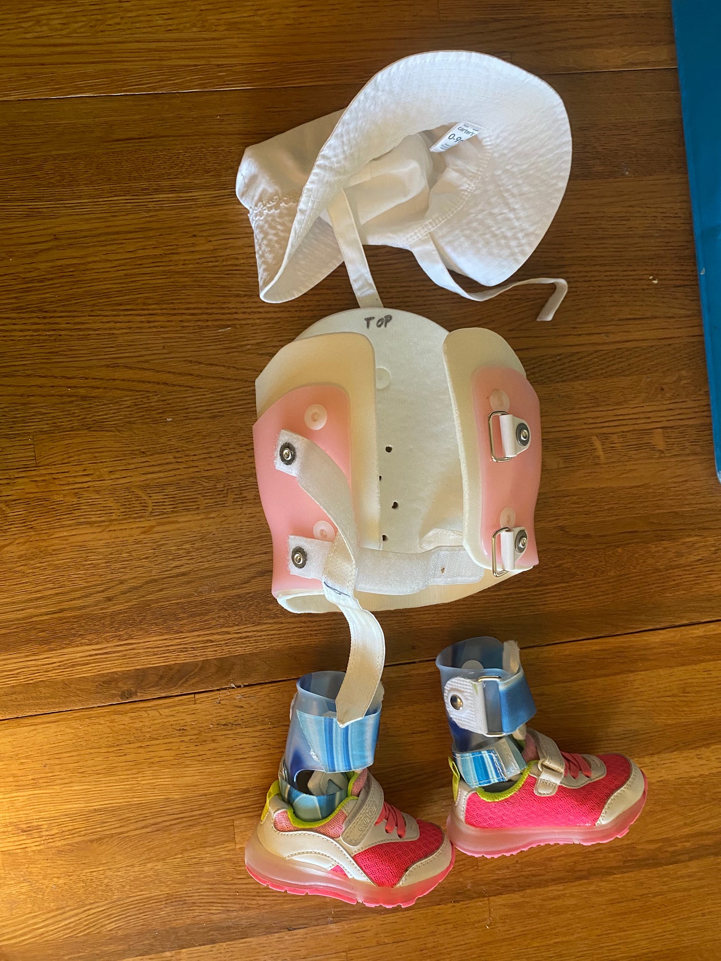 A baby-sized white cap, body cast in pink and blue AFOs with tiny pink sneakers lined up on a wood floor to look like an invisible child. 