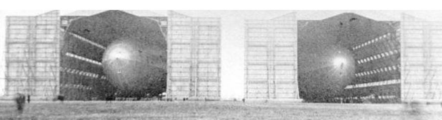 A grainy black and white photograph showing two large hangars with their doors open. Two Zeppelins are inside and we can see the curved blunt ends of both.