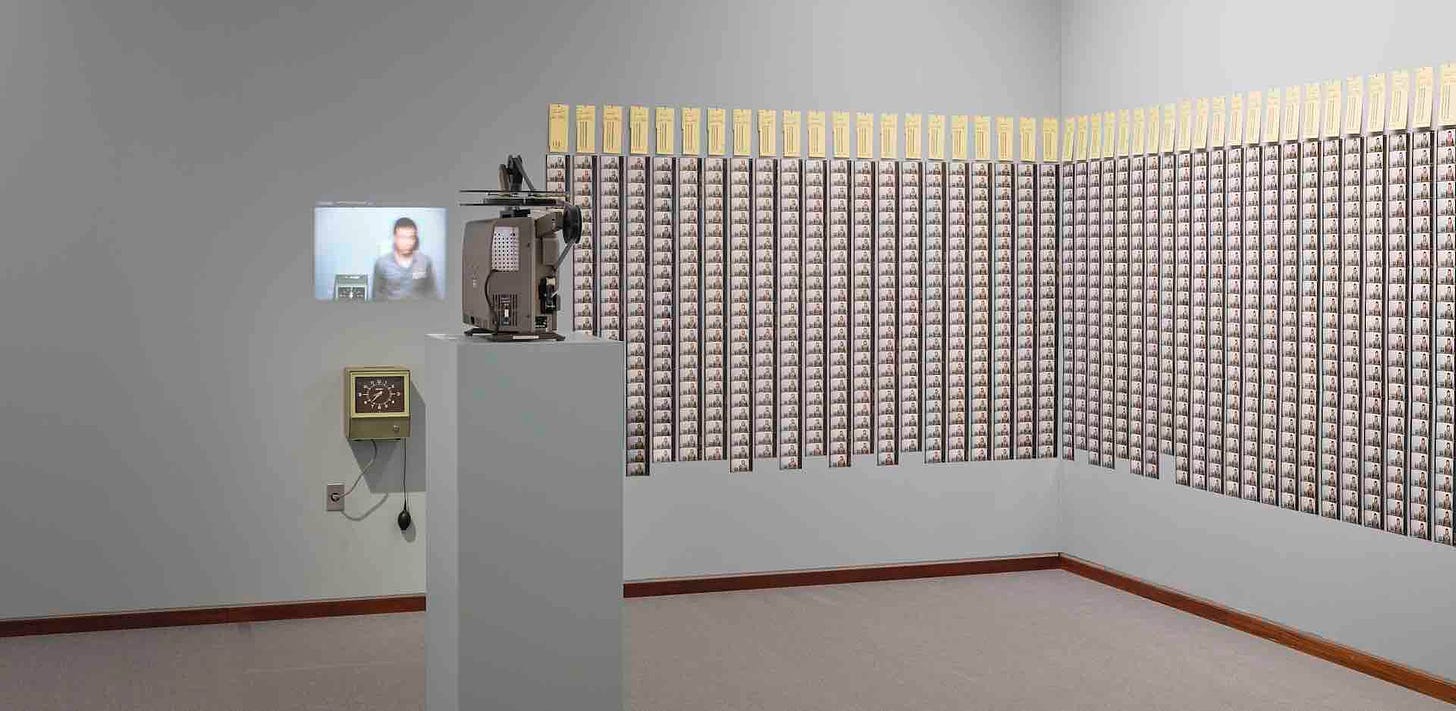 Imagem: Parede cinza com fotos polaroids coladas enfileiradas numa grande soma repetitiva. Vê-se também em cada fileira um cartão de ponto. Há na sala um projetor, o relógio de ponto e um filme sendo exibido na parede com imagem do artista.