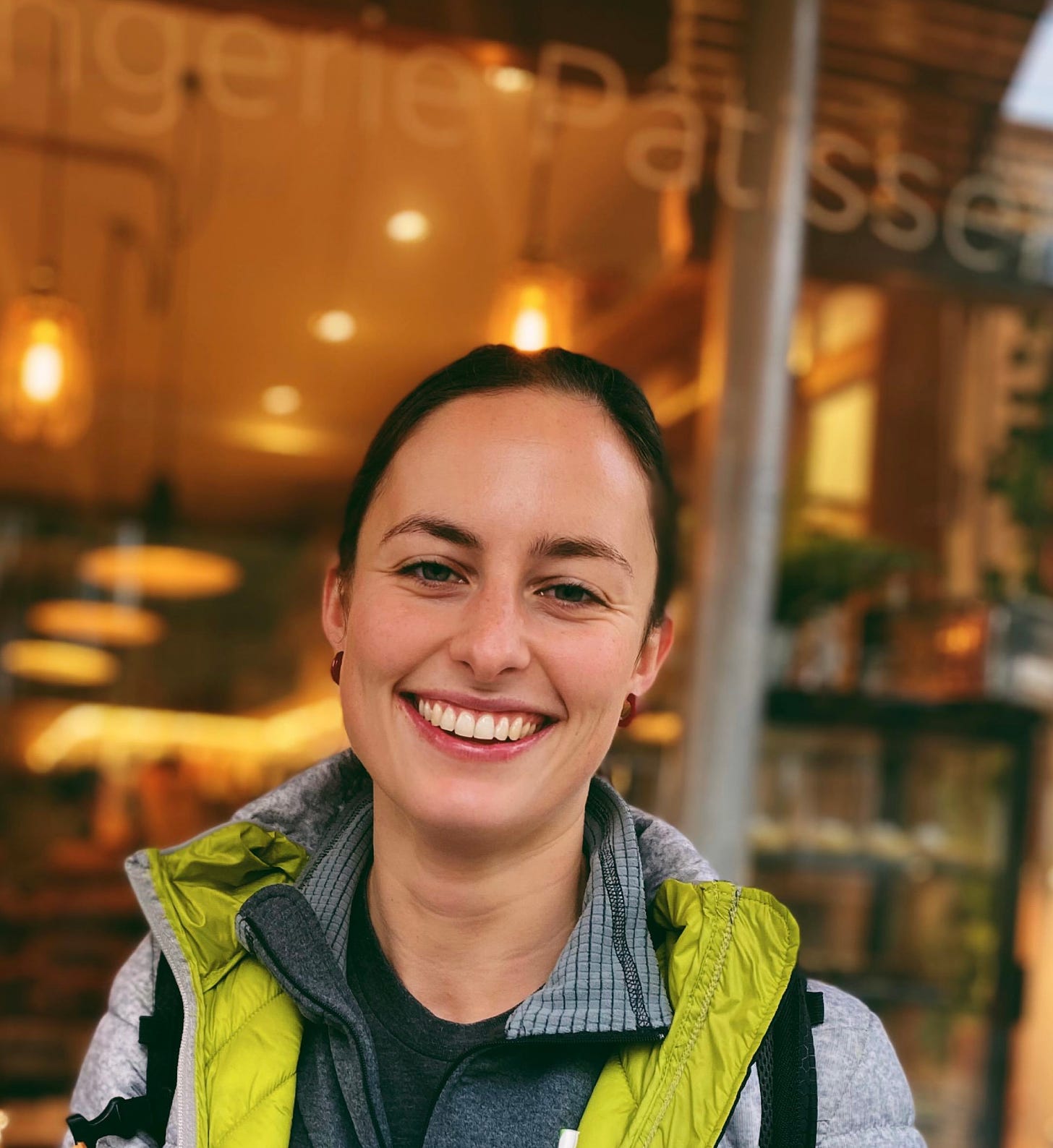 Chiara is a white woman with brown hai. In this photograph, her hair is tied back. She's smiling straight into the camera, her eyes squinting ever so slightly. She's wearing what appears to be a grey mountaineering jacket with lime-green lining.