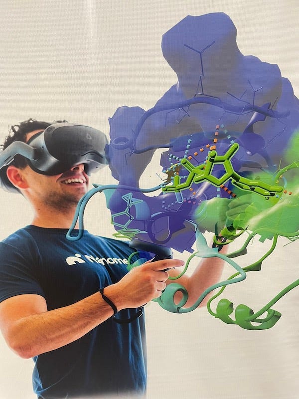 A man wearing virtual reality goggles and a t-shirt with the word ‘Nanome’ written on it looks up with a smile on is face. A multi-colored representation of a complex protein appears to be floating in the air above his upraised hands. A man wearing virtual reality goggles and a t-shirt with the word ‘Nanome’ written on it looks up with a smile on is face. A multi-colored representation of a complex protein appears to be floating in the air above his upraised hands.