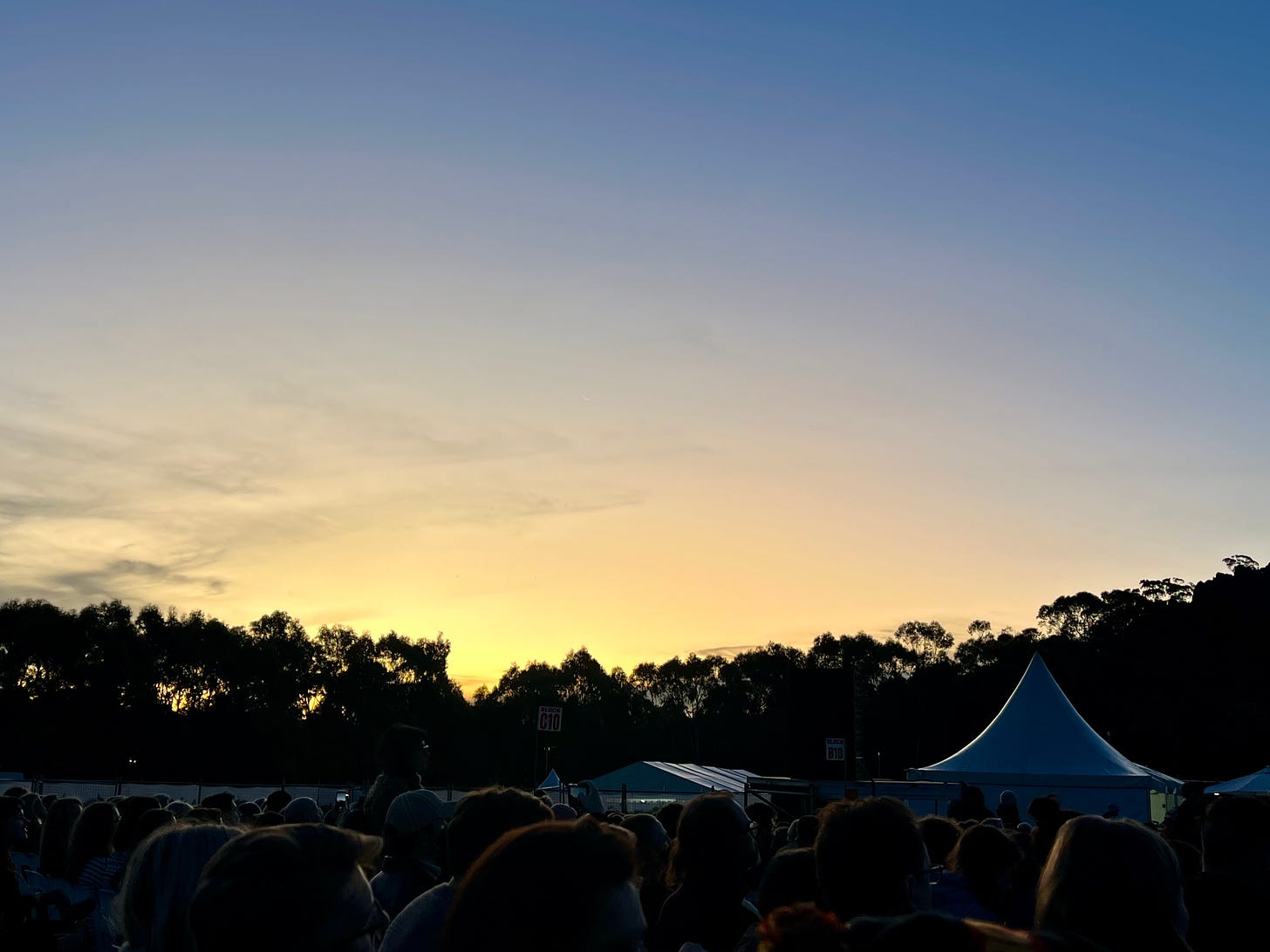 A sunset at a music festival. In the foreground, people and tents are backlit by a soft yellow glow. The sky is all pale yellow, orange, and blue.