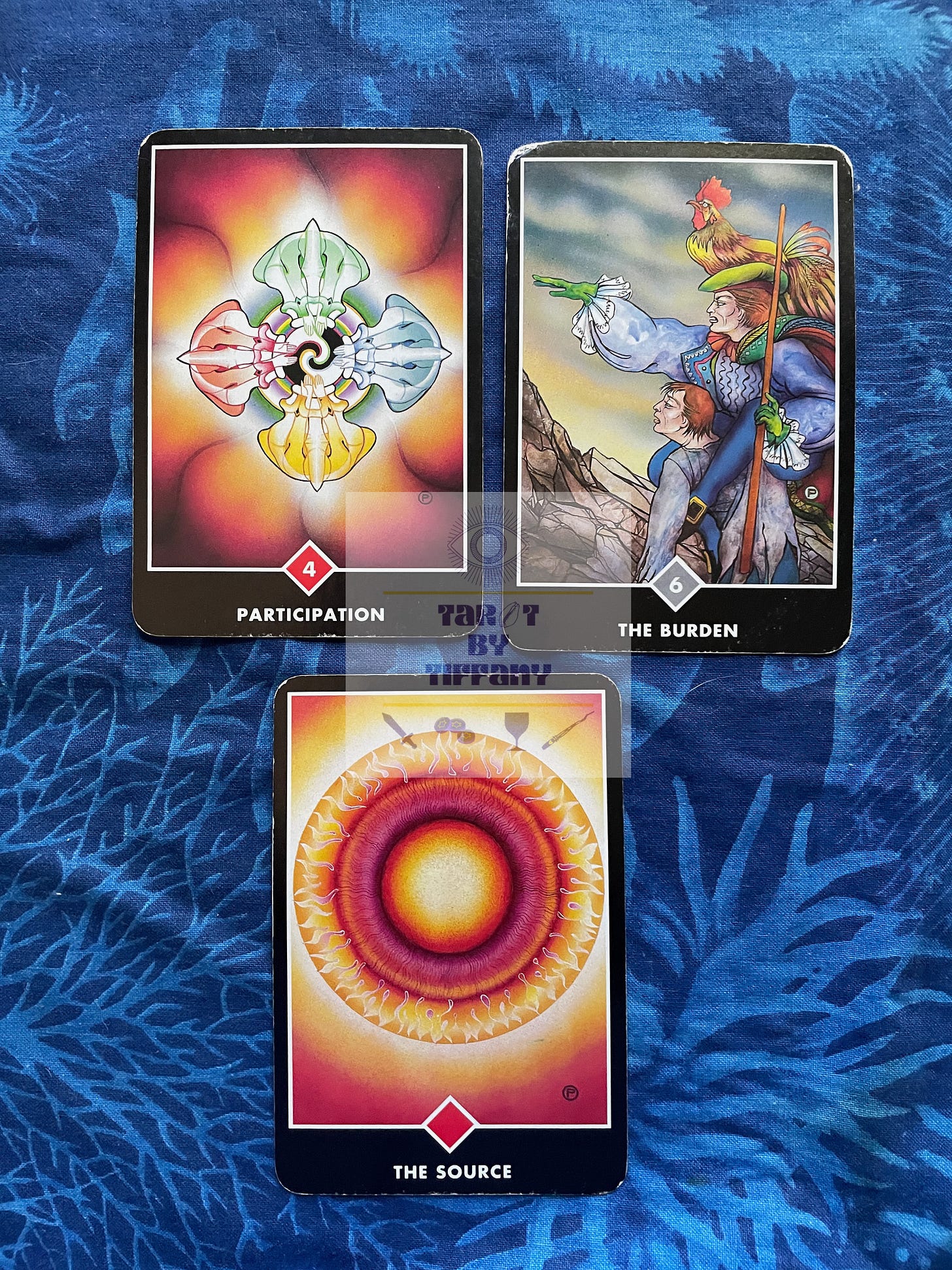 3-card reading with the Osho Zen Tarot. The cards are laid out on a cobalt blue cloth with an aquatic motif in light blue. Top two cards (L-R): 4 of Fire (Participation) and 6 of Clouds (Burden). Bottom card: Ace of Fire (The Source).