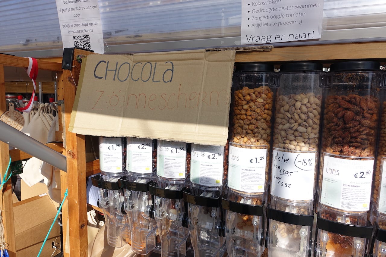 Over the containers containing chocolate goods, a cardboard sign has been tapped over the transparent container to stop them melting in the bright winter sunlight. On the cardboard, someone as written Chocola Zonnescherm