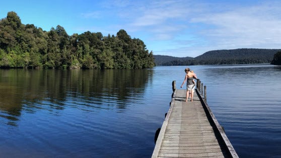 A person is walking along an old-fashioned jetty that extends into a lake. Native forest cloaks the hills behind and the sky and water are clear and blue.