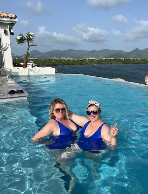 Amber Tamblyn and Eliza Clark pose together in a pool wearing matching dark blue one piece bathing suits. Amber has her arm around Eliza. They both wear sunglasses and smile broadly. Behind them, you can see the ocean and in the distance there are lush green hills.