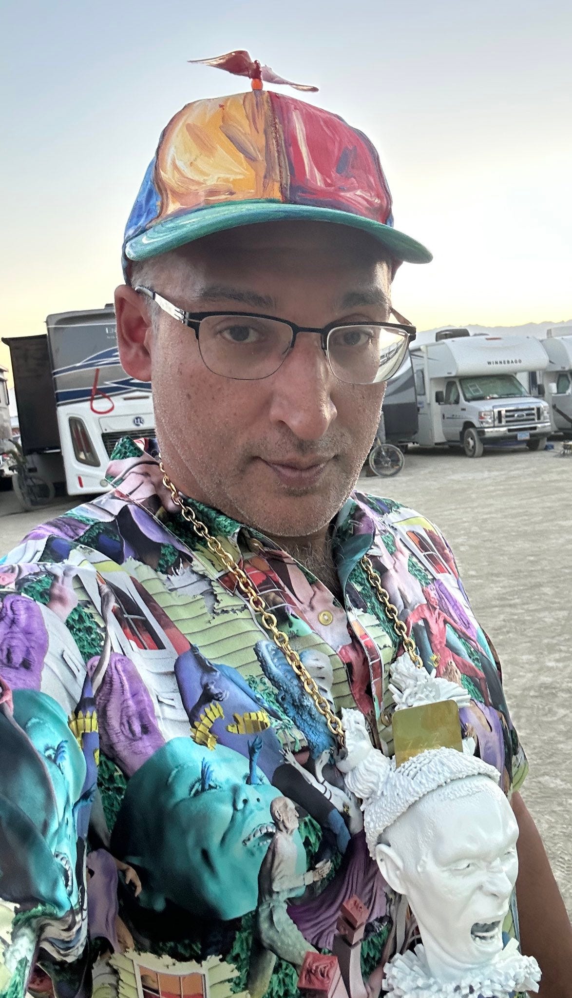 Selfie of Neal Katyal, a smug looking middle aged man wearing a paint-smeared style propellor beanie, an AI nightmare print shirt, and a gold chain with a large white bust of a screaming head (????) hanging from his neck. In the background is a line of dusty Winnebagos redolent of America’s most self-absorbed temporary desert RV park, Burning Man. His face looks like he’s about to say “My lover and I saw you from across the bar and we really dig your vibe.”