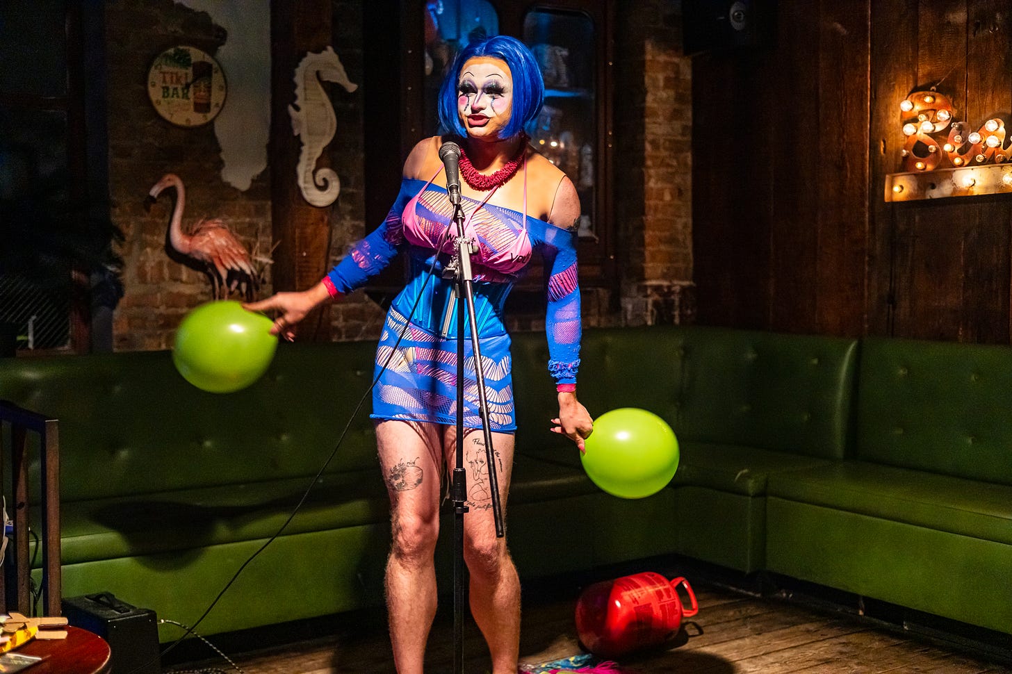 tank, a drag queen with three breasts in blue wig, pink bikini, blue mesh, corset, and clown makeup holds balloons and sings into mic at queers n peers variety show