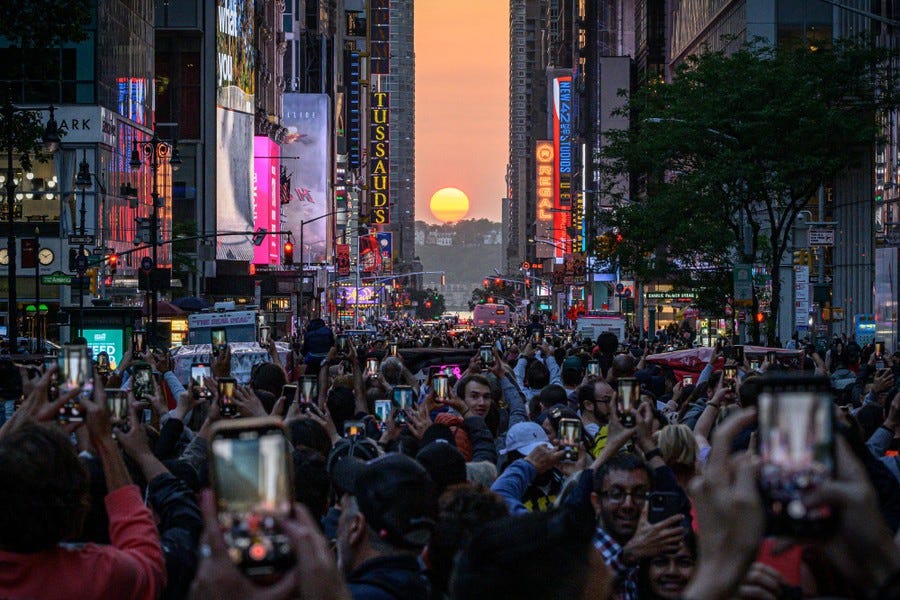 A city street is filled with people taking pictures of a sunset in the distance, between buildings