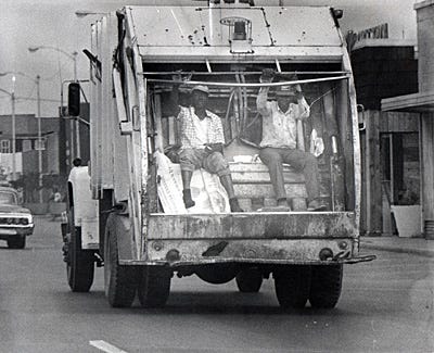 black and white photo of two Black sanitation workers sitting in the back of a moving garbage truck