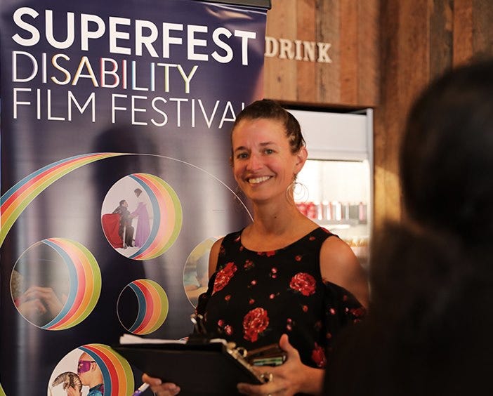 A photo of a white woman with her dark hair pulled back. She is wearing a black top with red roses on it, and standing in front of a disability film festival poster.