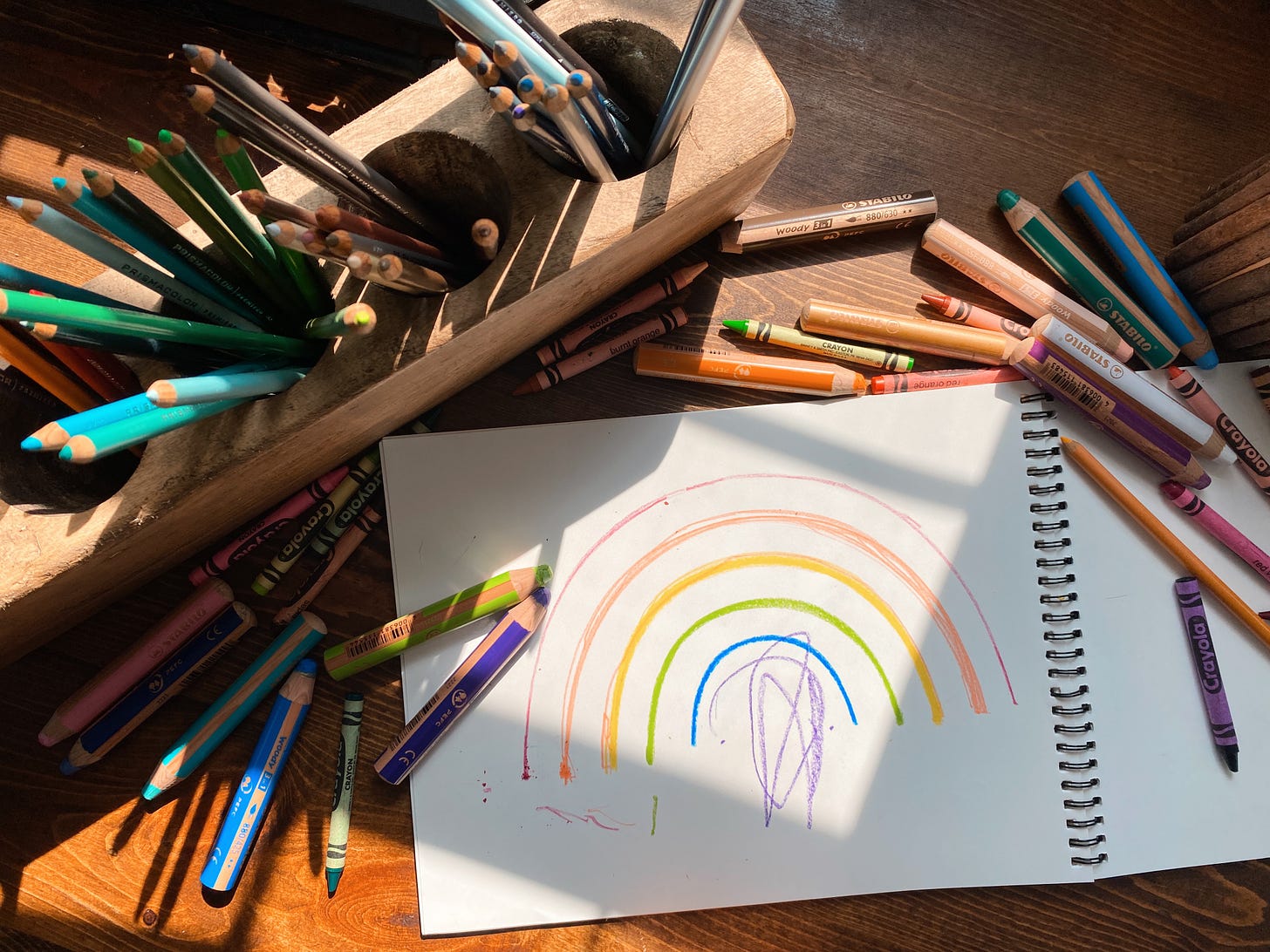 Crayons and colored pencils surround a sketchbook with a kid drawn rainbow. A shaft of sunlight comes in from the window.