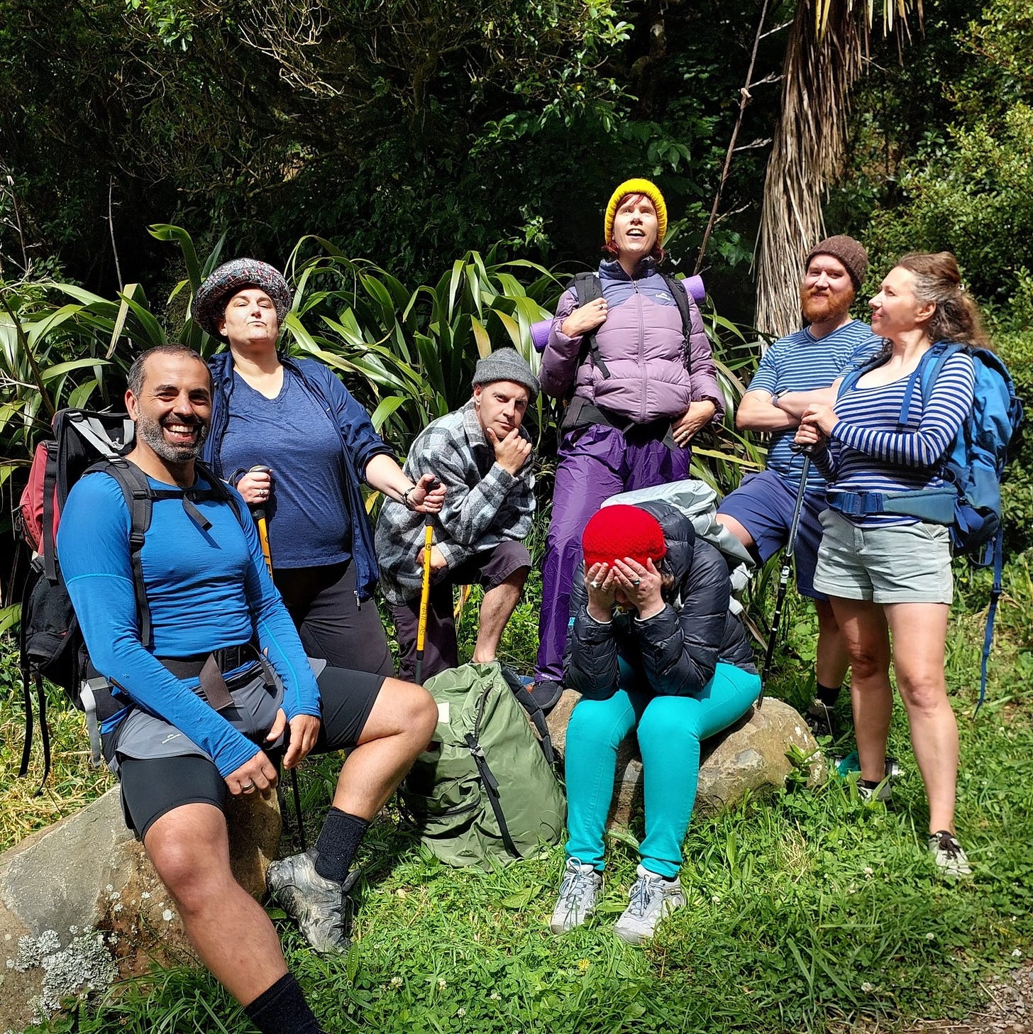 7 improvisors dressed ready to go tramping, with packs and thermals. Standing in a group outside by some flax bushes. They express a range of emotions.