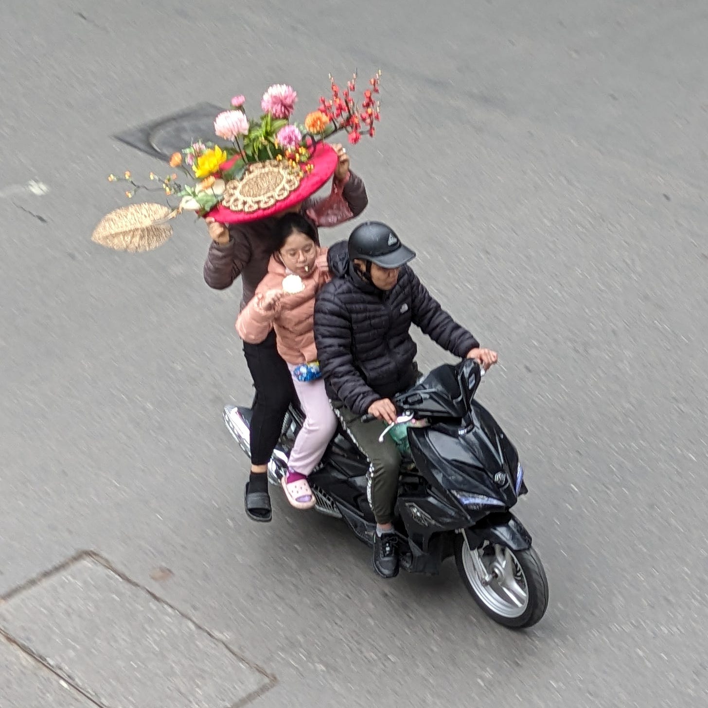 Three people on a scooter, father driving, mother in the back holding a large decorative platter above her head with both hands, daughter in the middle, both hands occupied with her snack