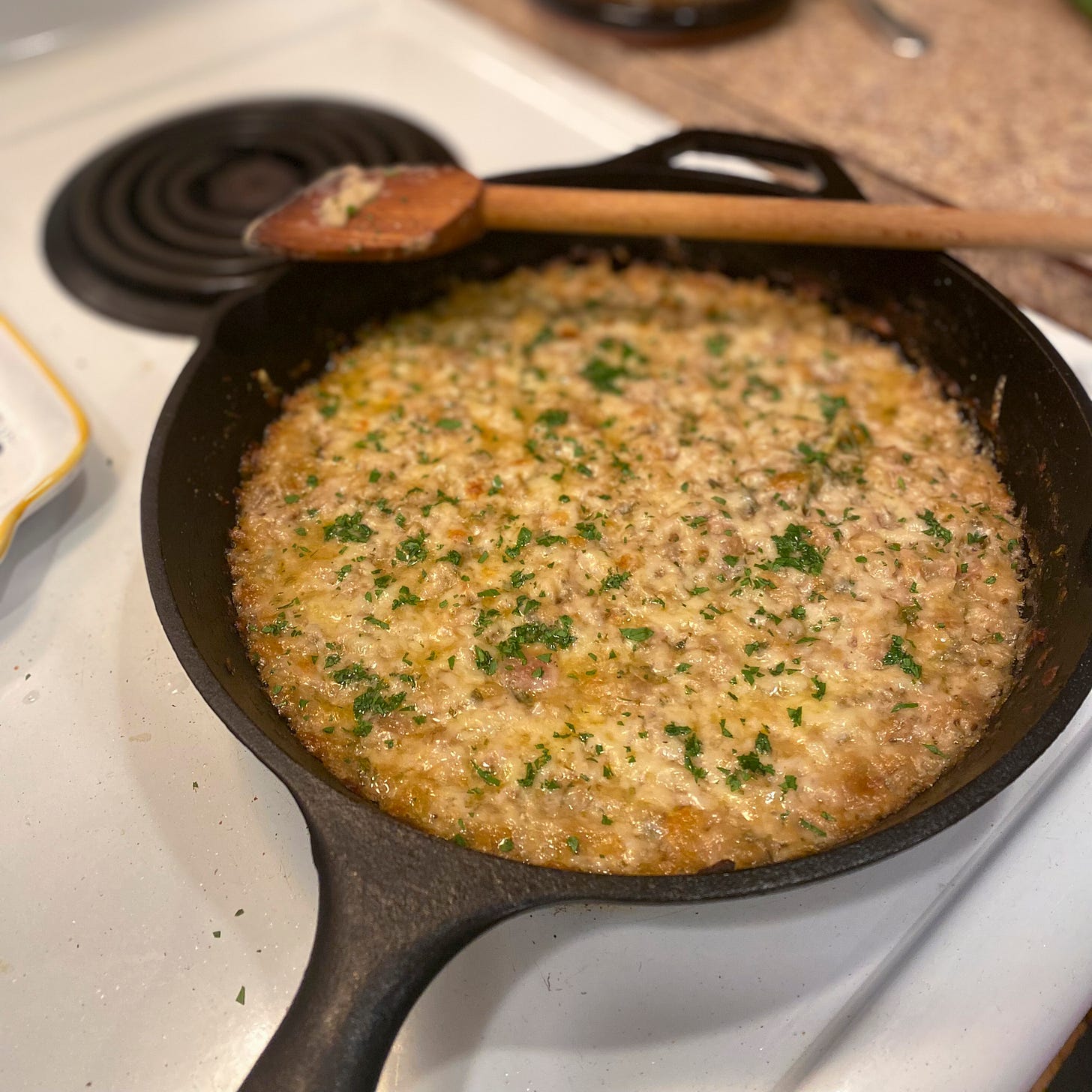 A cast iron pan with a wooden spoon resting on its edge, the pan full of the tuna white bean casserole described above. Cheese is melted and browned on top and it's dusted with chopped parsley.