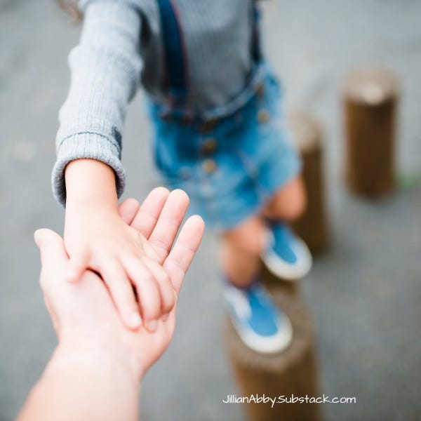 A child reaches out their hand to an adult's for support as they step across wooden posts.