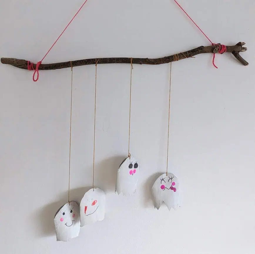 the image shows a cute ghost craft for kids. 4 ghosts made from toilet paper tubes have been turned into a mobile that have been hung from a branch using yarn. 