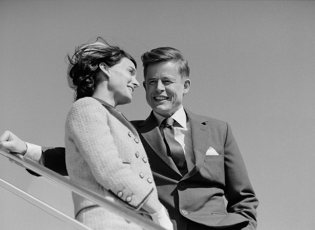A man and a woman stand on a plane's stairways. It is in black and white. The woman is looking towards the man. The man wears a suit and is looking towards the woman.