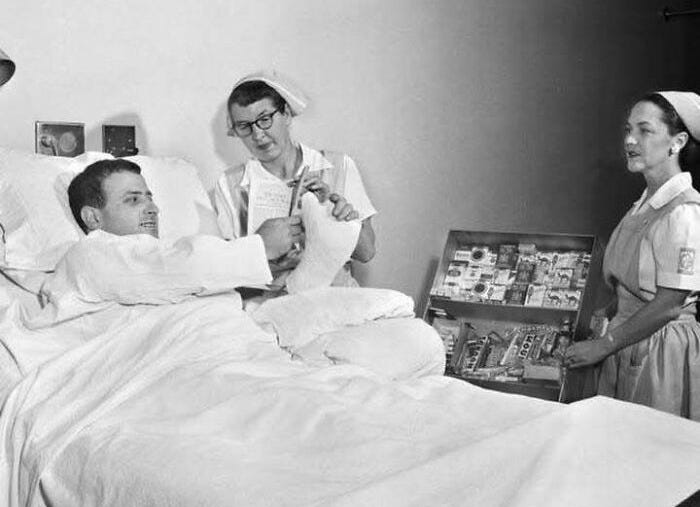 Black and white photo of a patient with a cast in a hospital bed being attended by two nurses, showing past medical care.