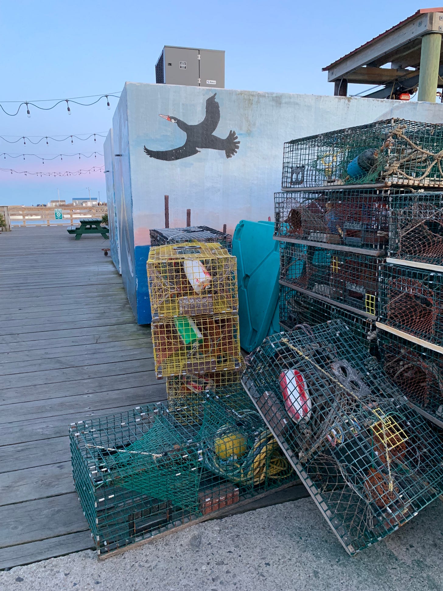 A mural of a bird flying at dusk in front of actual dusk which is identically colored. There are a bunch of lobster traps in the foreground, yellow and green.