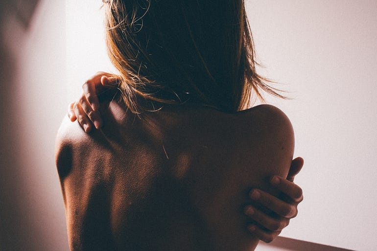 Picture of a thin woman’s nude back. She has her arms wrapped around herself. She could be hugging herself or trying to scratch an itch on her back.
