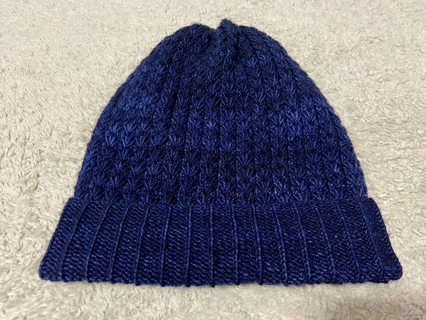 The Constellate Hat with a folded brim is hand-knitted with dark blue yarn and resting on a cream-colored throw.