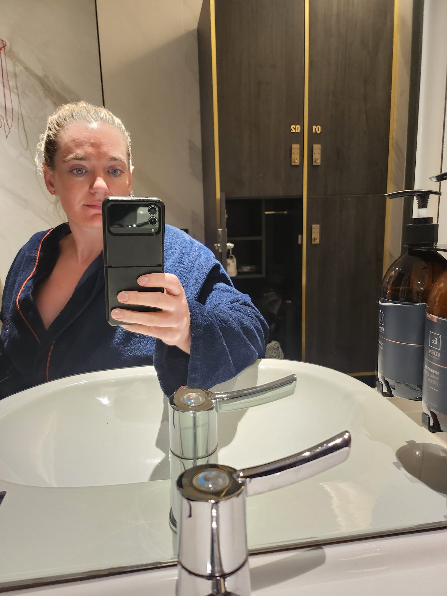 I take a selfie in a bathroom mirror. Wearing a navy robe with gold lining, this is Eden One. The branding can be seen on the labels of the soap bottles in the mirror. Lockers are behind me. 