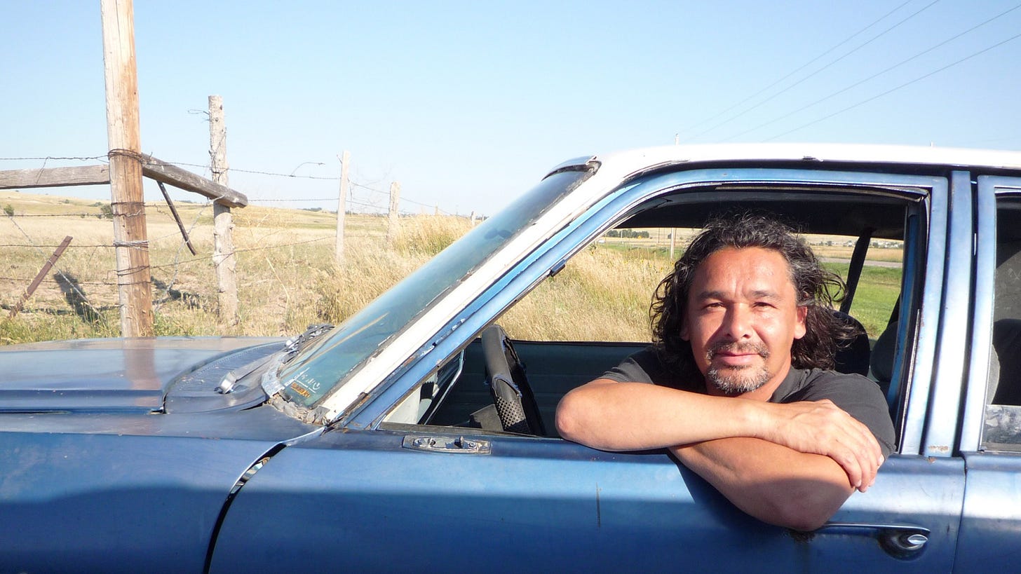 This image shows Cree-Canadian filmmaker Neil Diamond, director and narrator of the 2009 documentary titled Reel Injun: On the Trail of the Hollywood Indian, sitting in the blue "rez car" that he drives across the United States while investigating how Hollywood films have fixed stereotypes of Native Americans in the minds of global audiences. Diamond looks out his car's driver's-side window directly into the camera. The car sits in front of a fenced field somewhere in the American Midwest or West.